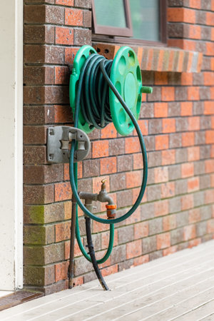 Watering garden equipment. Old garden hose, reel and adjustable spray gun, pistol nozzle. Dirty old garden hose reel mounted on the back of old brick house.の写真素材