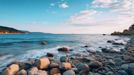An evocative scene ideal for travel brochures, nature-themed articles, or as calming wall art | This image exquisitely captures the tranquil beauty of a twilight seascape, where the gentle motion of the sea meets a rugged shoreline strewn with smooth, round stones. The soft light of early evening casts a warm hue across the scene, imbuing the composition with a sense of peace and stillness. The long exposure creates a silky effect on the water's surface, blurring the waves and lending an ethereal quality to the scene that contrasts with the solidity of the stones. In the background, the coastline extends into the distance, suggesting the vastness of nature and the serene isolation one can find there. This visual narrative is punctuated by the clear blue sky, with subtle wisps of clouds and contrails that draw the eye upward and convey a feeling of expansive freedom | Perfect for environmental publications to illustrate the serene beauty of coastal regions, as an element of design in travel agency offers promoting seaside getaways, or as part of a collection in a gallery focused on natural landscapes. It serves as a peaceful visual escape in interior design, providing viewers with a sense of calm and spaciousness, or as a thoughtful backdrop in wellness and meditation spaces | a landscape photograph showing casing a serene inviting coastal environment during the golden hour, with a play of light and shadow over the water and stones, offering depth and texture while contemplation, and leaving plenty of room for overlaying inspirational messages or branding elementsの素材