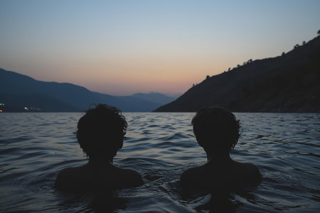 An evocative image perfect for travel blogs, lifestyle magazines, or introspective editorial pieces | This serene photograph portrays two individuals submerged in water up to their shoulders, against the canvas of a waning twilight sky. The silhouetted figures convey a sense of peaceful companionship, while the subtle ripples around them capture the gentle movement of the water in an otherwise still environment. The harmonious blend of navy blues and dim oranges in the sky above suggest the concluding moments of sunset, a transition from day to night that inspires reflection. The layered mountains in the background deliver a majestic sense of scale and the progression of natural beauty fading into the distance. This image's composition, with its strong horizon line and the placement of the figures in the lower third, evokes tranquility and the simplicity of a moment shared between people within the vastness of nature | Ideal for use in content that discusses mental health, the beauty of everyday moments, or the benefits of connecting with nature. The photograph could also serve in marketing materials for romantic or wellness getaways, and as a poignant visual metaphor in narratives about friendship, contemplation, or the intersection of human and natural beauty | a silhouette of two people enjoying a calm swim with a pastel-hued sunset and mountain silhouette in the background, offering a sense of tranquility and intimacy amidst the grandeur of the landscape, suitable for artistic overlay or standalone useの素材