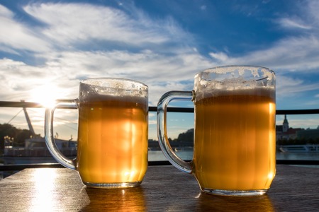 Close-up two cold beers with foam and water drops on the background blue sky and white clouds and the sunの写真素材