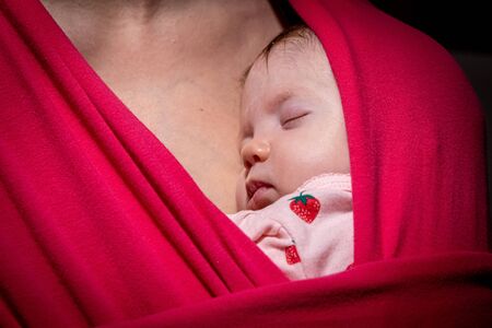 Mother carrying her cute baby daughter in sling. Newborn baby sleeping in a sling, in the embrace of her mother. The baby girl is 2 months old.の写真素材