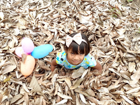 Ranong/Thailand- 3.20.2020: A cheerful asian girl, her sitting pose on dry leaves in summer and staring at the camera, with multicolored balloons under, the natural background of brown leafs concept.のeditorial素材