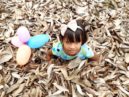 Ranong/ Thailand- 3.20.2020: A cheerful asian girl, her sitting pose on dry leaves in summer and staring at the camera, with multicolored balloons under, the natural background of brown leafs concept.のeditorial素材