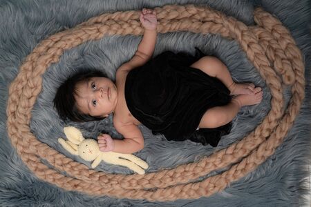 Full body picture of the boy lying on a grey fur having a rabbit toy, covering with a black blanket looking curious at the viewer.の写真素材