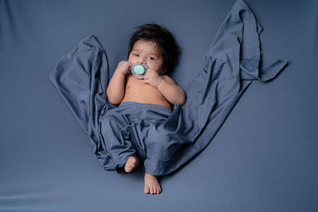 Portrait of baby boy with pacifier leaning on the bed covering by blue shawlの写真素材