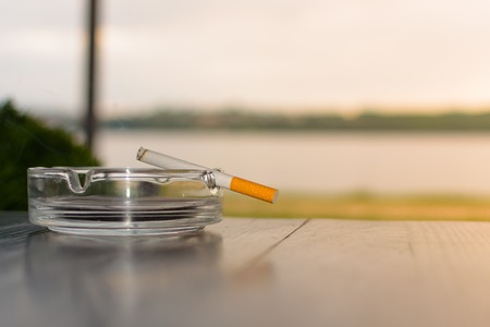 Burning cigarette in the ashtray on the wooden table against the background of the lake