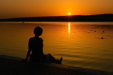 The girl sits on the shore of a pool in the city and looks at the sunset.の写真素材