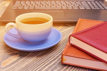 A cup of tea and two notebooks with a laptop on a wooden table. Side view. Small depth of field.の写真素材