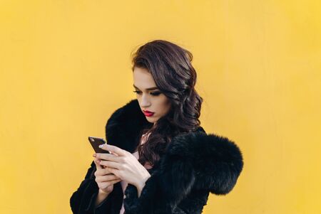 Trendy beautiful cool hipster blond girl wearing a gray hat and black leather jacket taking selfie with mobile phone against a yellow wall. Making peace hand gesture and smiling.の写真素材