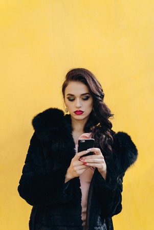 Trendy beautiful cool hipster blond girl wearing a gray hat and black leather jacket taking selfie with mobile phone against a yellow wall. Making peace hand gesture and smiling.の写真素材