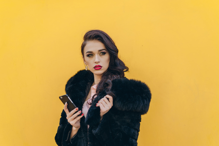 Trendy beautiful cool hipster blond girl wearing a gray hat and black leather jacket taking selfie with mobile phone against a yellow wall. Making peace hand gesture and smiling.の写真素材