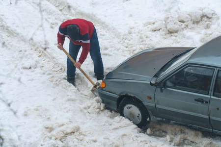 A man digs a shovel of snow from a car stuck in the snowの写真素材