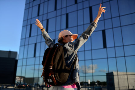 A happy traveler woman with a pink hat and a black leather backpack enjoys a stunning view of the city downtown with raised hands.の写真素材