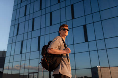 Portrait of handsome tourist hipster man in sunglasses with bag handy, standing guy near the building in downtown. Calm relaxing moment, summer vacation conceptの写真素材