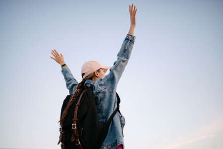 Attractive girl standing with raised hands against blue sky empty background. A happy traveler woman with a pink hat and a black backpack enjoys a stunning view of the city with raised hands.の写真素材