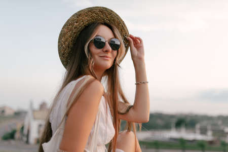 Portrait of a relaxed happy woman in white dress with hat looking at the camera. Cityscape in the backgroundの写真素材