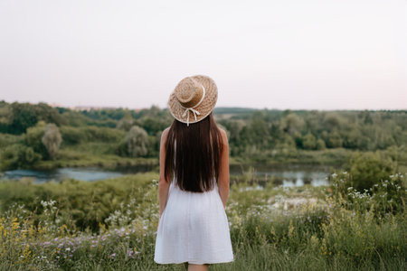 A serene, beautiful view of a woman wearing a flowing white dress and straw hat, gazing at a...