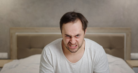 A frustrated man is expressing his deep anger and emotions while sitting in his bedroomの写真素材