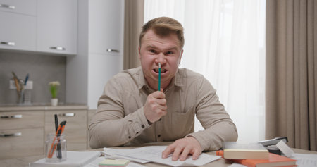 Frustrated Male Working from Home Surrounded by Papers and Holding a Pen in Hand