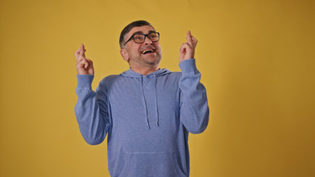 Hopeful man in his 50s with crossed fingers on yellow background