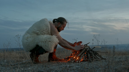 Man in animal hide kneeling and blowing at burning campfire against cloudy sky in evening in...
