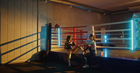 wo young athletic women preparing for sparring in a boxing ring with colored lighting in the gym,...