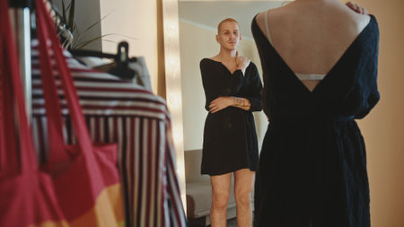 A guy at home, gay, standing near a lighted mirror wearing a womans dress and watching in the mirror