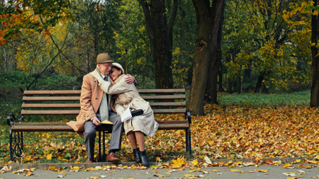 Elderly couple sitting reading a letter together on a bench in an autumn park then smiling and...