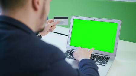 A young man using a laptop with a green screen and holding a credit card indoors. Close-up shot....