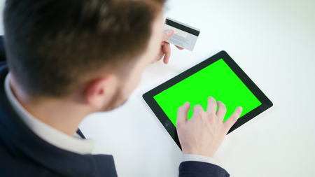A young man using a tablet with a green screen and holding a credit card indoors.View from the top. Close-up shot. Soft focus.の写真素材