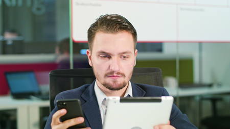 A young businessman using a smartphone and a tablet in the office. Close-up. Dolly shot. Soft focus.