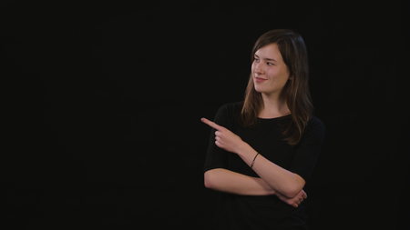 A beautiful young lady smiling and standing with her arms folded and pointing with her finger against a black background. Medium Shotの写真素材