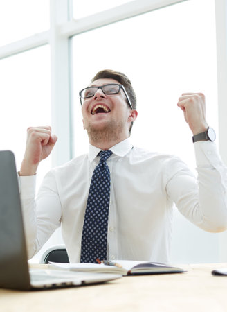 Happy young businessman raising hands in front of laptop at office deskの写真素材
