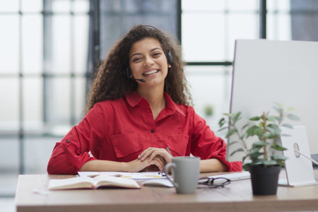 A young attractive woman in a red shirt sits at a table in a red jacket in headphones with a microphone.の写真素材