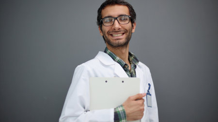Portrait of positive male doctor posing and smiling at camera on gray studio backgroundの写真素材