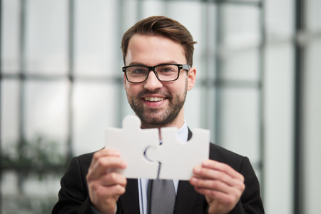 Portrait of optimistic bearded businessman holding puzzle pieces, solving tasks, looking at cameraの写真素材