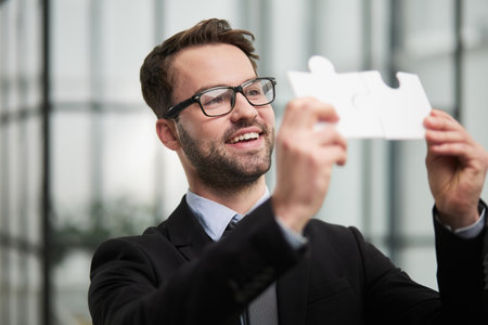 Portrait of optimistic bearded businessman holding puzzle pieces, solving tasks, looking at cameraの写真素材