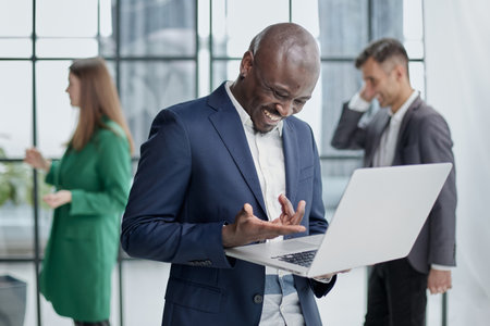 Young african american man in business suit holding laptop while standing at office corridor