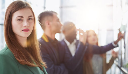 Thoughtful woman looking at camera while colleagues in the background at sticky notes on a glass wall.の写真素材