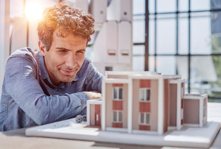 Young Male Designer Assembling Wooden Pieces into a House Miniature on Top of the Table with Blueprint.の写真素材