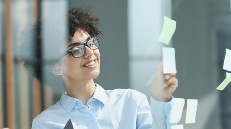 a girl in a modern office behind glass uses sticky notes and makes notesの写真素材