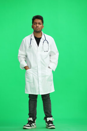 A full-length male doctor on a green background holds his hands in his pockets