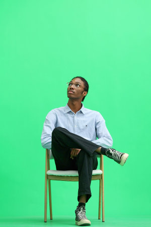 A man in a gray shirt, on a green background, is sitting on a chair