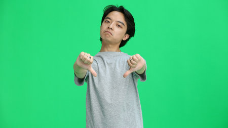 A guy in a gray T-shirt, on a green background, close-up, shows his thumbs downの写真素材