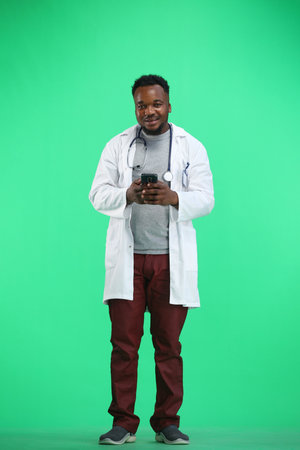 The doctor, in full height, on a green background, uses a phoneの写真素材