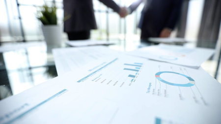 Two people shaking hands in front of a table with papers on it. The papers are spread out and have numbers and graphs on them. Scene is professional and seriousの写真素材