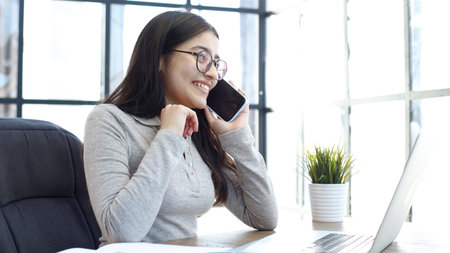 A young woman with glasses in the office is working on a laptop and talking on the phoneの写真素材