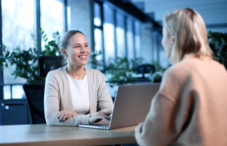 Two women are sitting at a desk with a laptop in front of themの写真素材