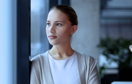 A woman is standing in front of a window with her hand on her chinの写真素材