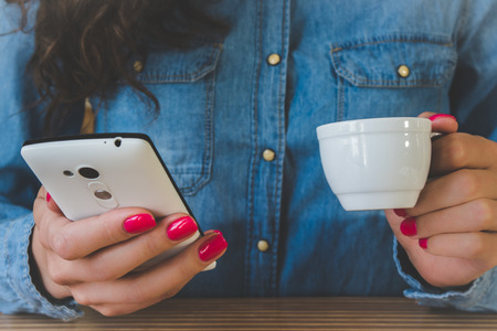 The girl in the blue denim shirt sitting at the table. In one hand a white Cup of espresso, and the other with a white mobile phone. Red manicure. Vintage photo.の写真素材